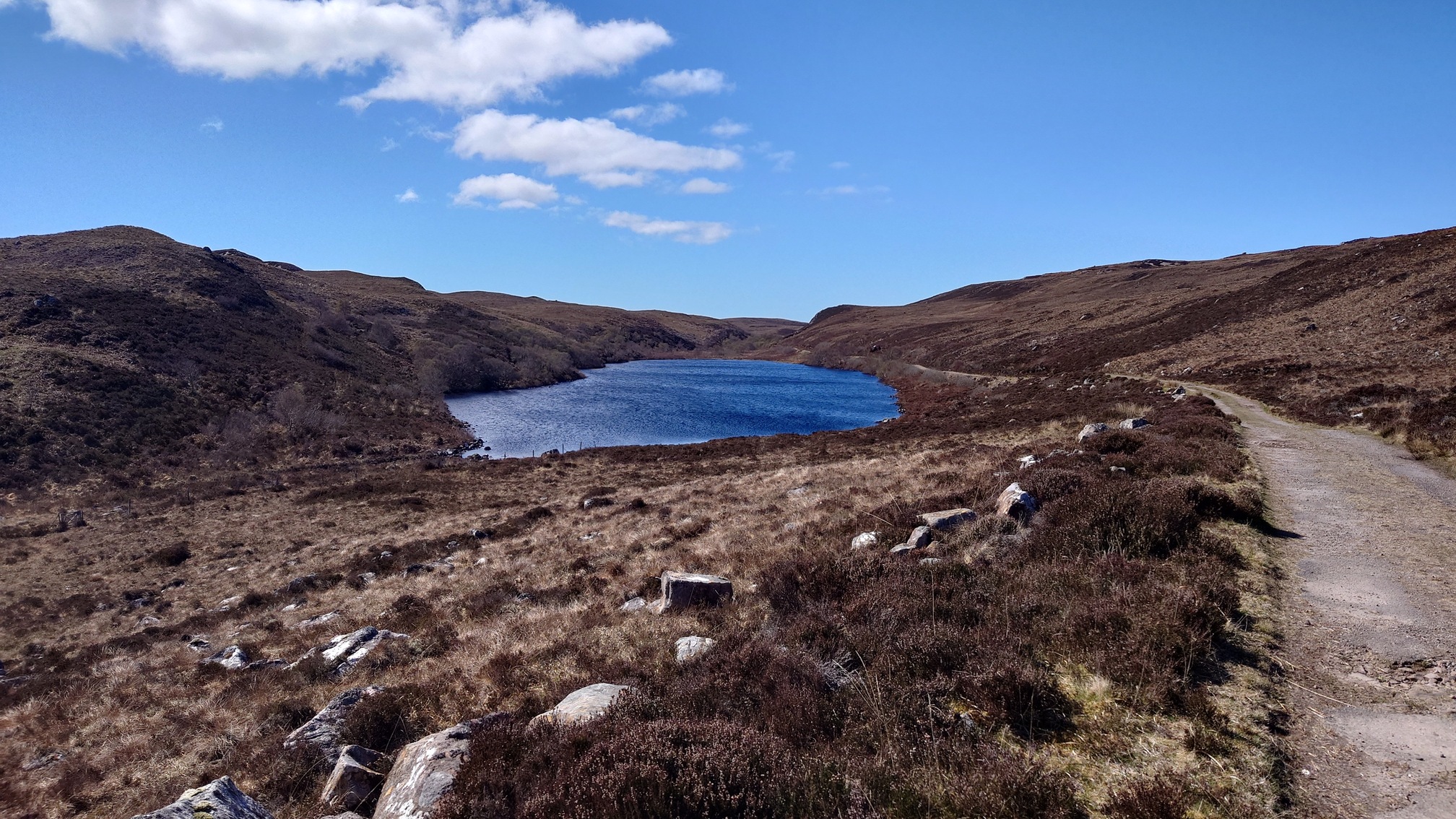 An old road curls through heather and grass past a small blue loch