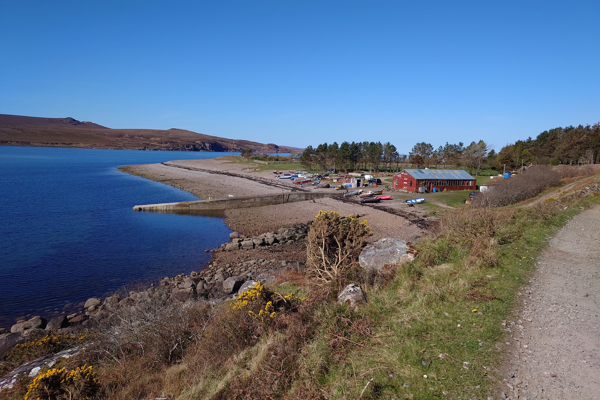 Looking down at the landing site, a rocky beach with a concrete jetty. A dozen or more small boats are hauled up above the high water, and around a large red wooden building are lots of piles of materials. There are trees behind the building.