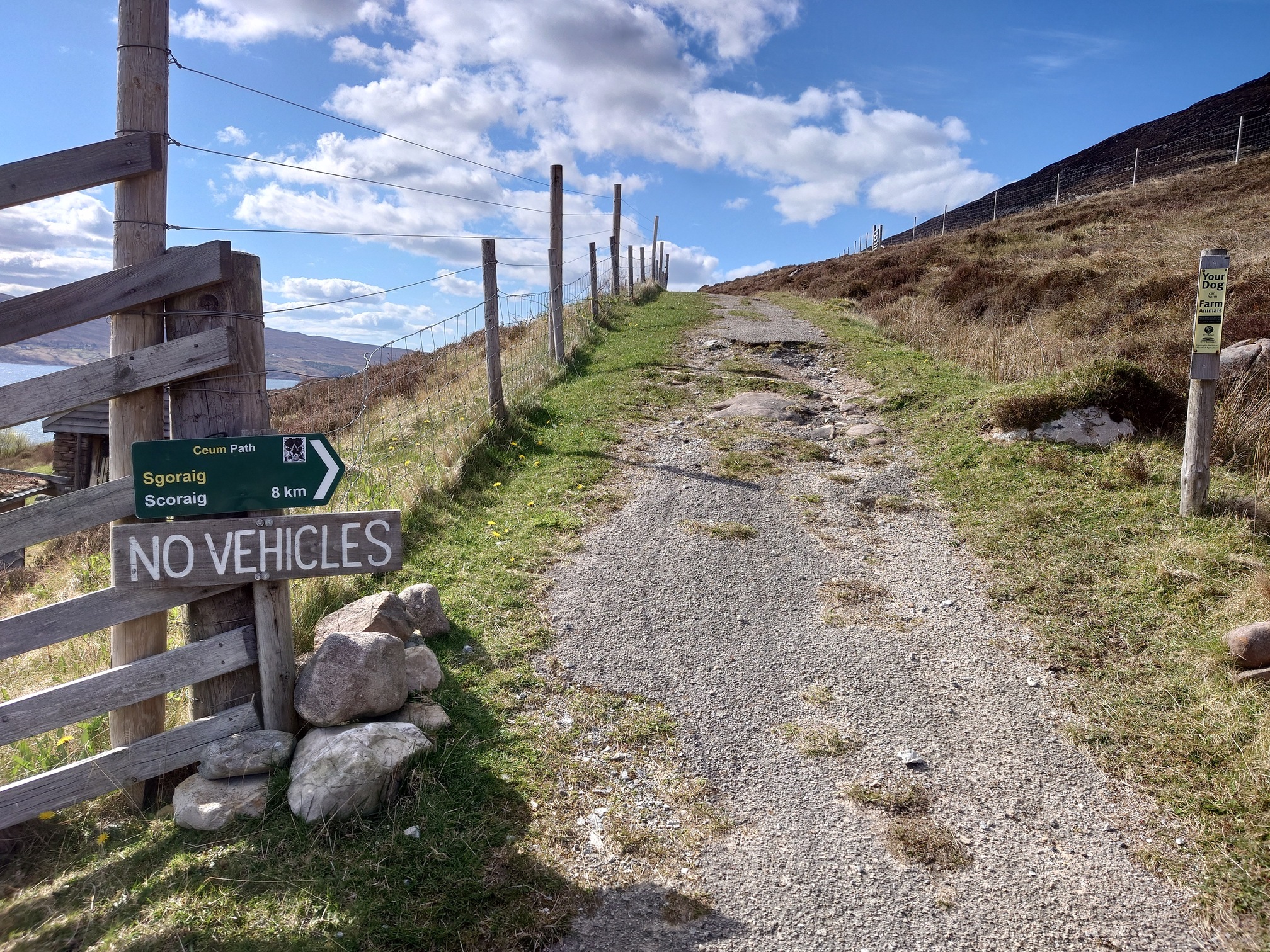 An old tarmac road, worn and grassy, leads past a green sign pointing the way to Scoraig in English and Gaelic. A hand painted sign reads NO VEHICLES
