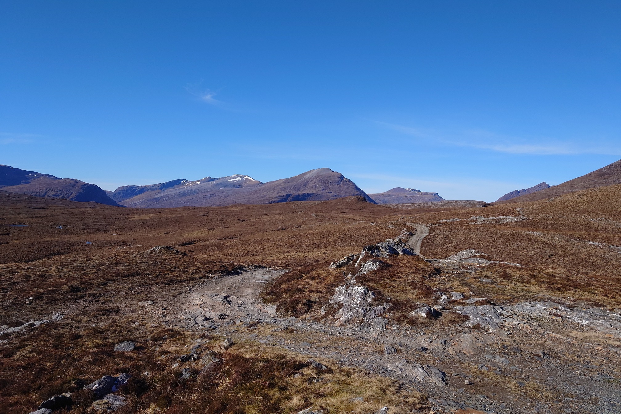 A rocky path stretches across barren moorland towards a mountainous horizon under a clear blue sky. Some of the mountains have patches of snow and theres a few wispy clouds in the sky