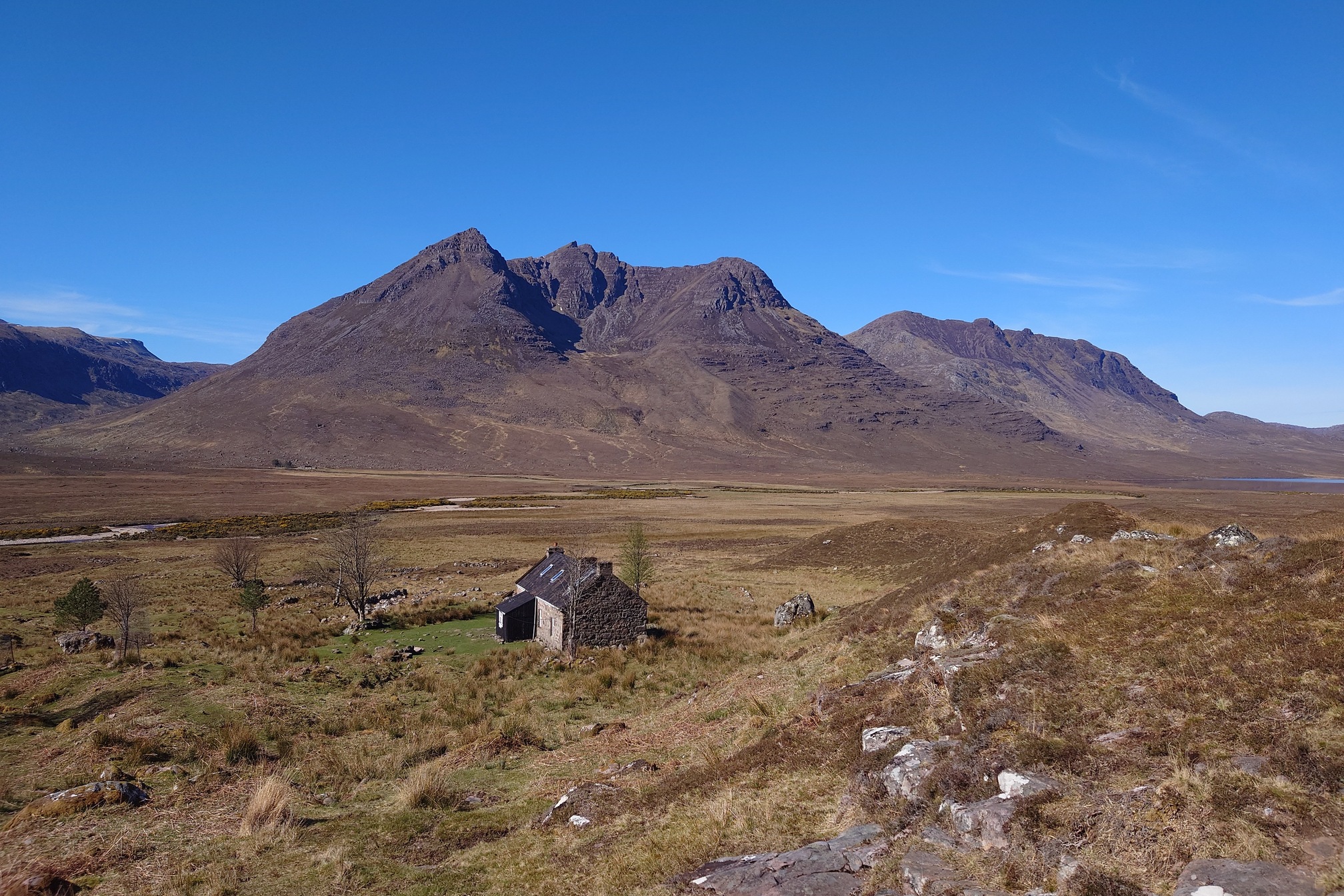 a small one story cottage, Shenavall bothy. There are a few scraggly trees around it, and it sits at an epic location with the mountain Beinn Dearg Mhor behind.