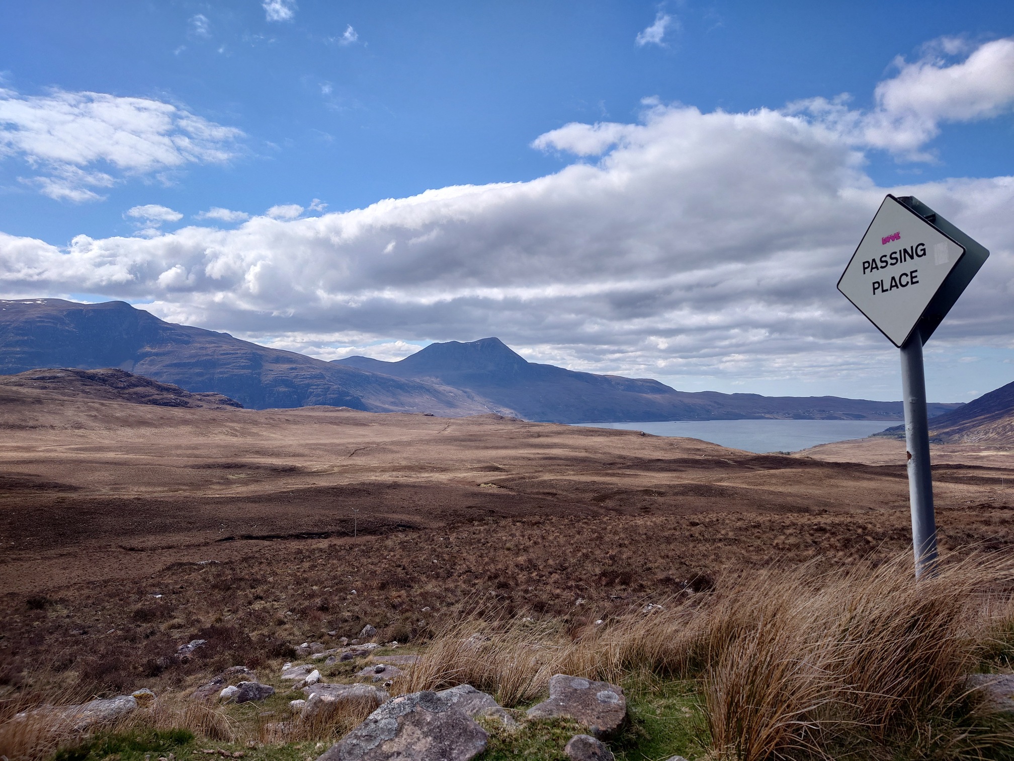 A white diamond Passing Place sign stands in the grass. Behind it is a sunlit vista of Scottish uplands, the ground brown with heather and dry grass, stretching to distant mountains and a loch under a blue sky