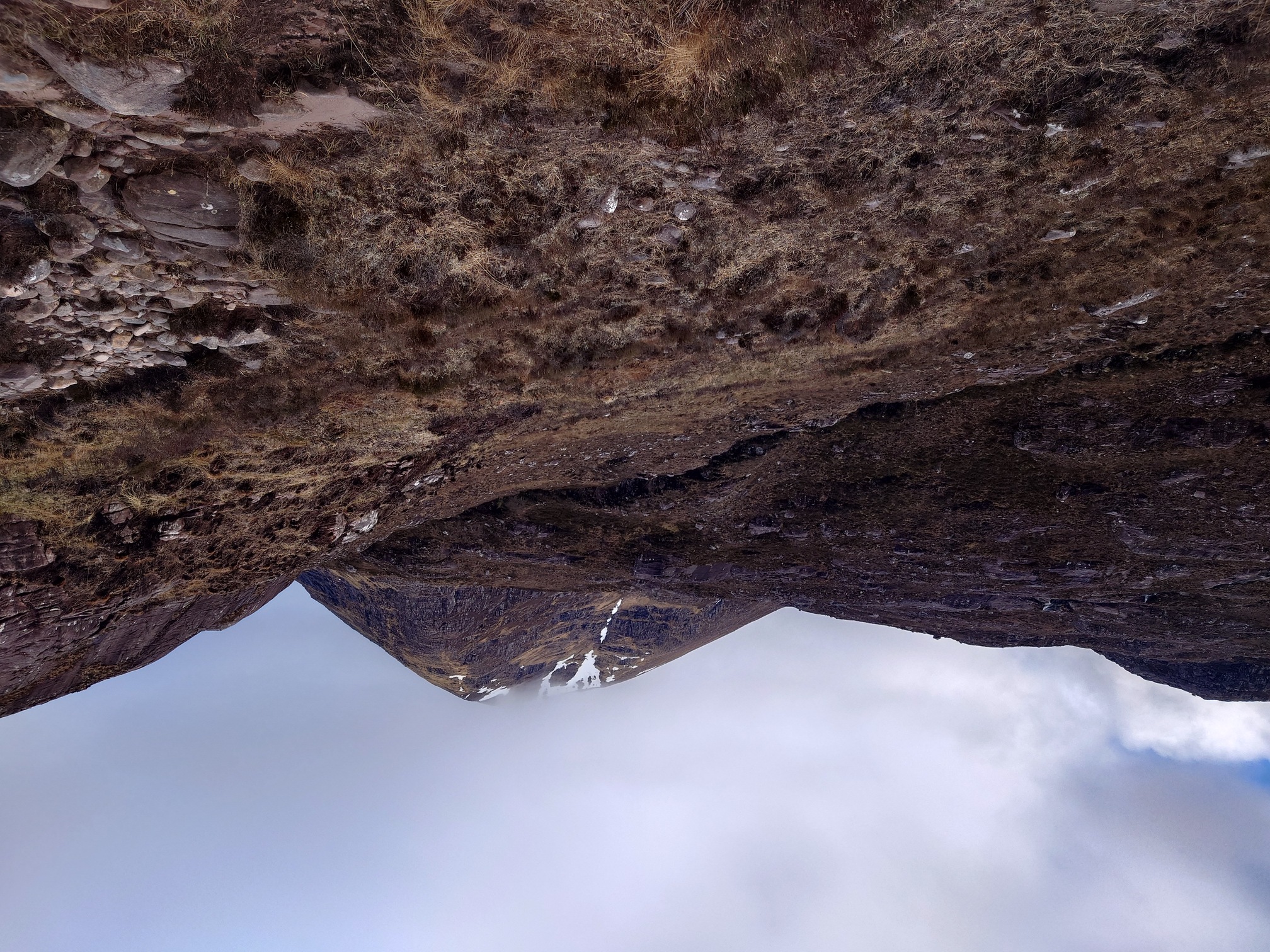 rocky ground with grass and heather and the gully of a stream lead very steeply uphill towards a pyramid-chaped mountain, the top dappled with snow and the very peak in the cloud