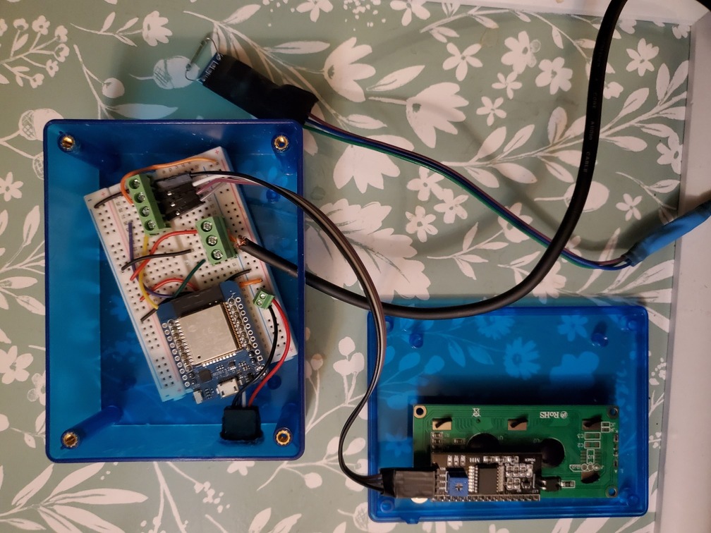 A translucent blue box containing a small form ESP32 on a solderless breadboard, with various wires and connectors. the back of the lid has the LCD unit connected to it.